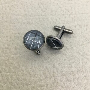 May include: A pair of round cufflinks with a gray and white grid pattern. The cufflinks have a silver-colored metal backing and post. The design is a geometric pattern of white lines on a gray background.
