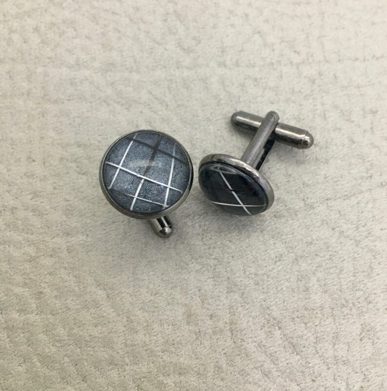 May include: A pair of round cufflinks with a gray and white grid pattern. The cufflinks have a silver-colored metal backing and post. The design is a geometric pattern of white lines on a gray background.