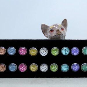 May include: A collection of round stud earrings with various colorful abstract designs. Each earring has a silver-colored bezel. The designs include white, black, pink, blue, and green lines on a colored background. A ceramic cat figurine is in the background.