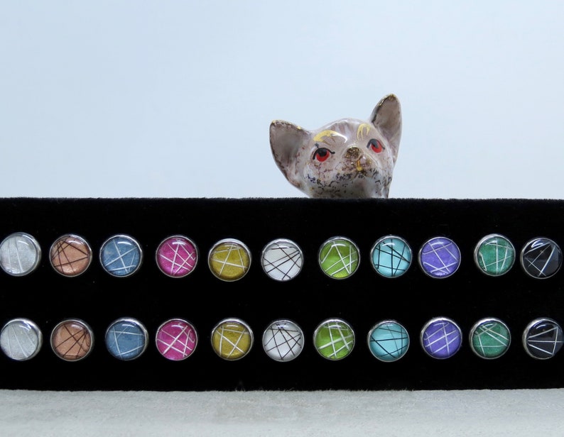 May include: A collection of round stud earrings with various colorful abstract designs. Each earring has a silver-colored bezel. The designs include white, black, pink, blue, and green lines on a colored background. A ceramic cat figurine is in the background.