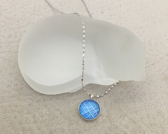 Puede incluir: Un collar de cadena de plata con un pequeño colgante redondo. El colgante es azul con un patrón de cuadrícula blanca.