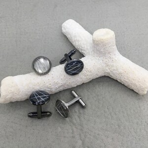 May include: Four pairs of cufflinks with a circular design. The cufflinks have a black base with a white, linear pattern. Two pairs have black stems, and two have silver stems. The cufflinks are displayed on a white, coral-like object.