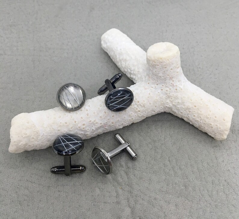 May include: Four pairs of cufflinks with a circular design. The cufflinks have a black base with a white, linear pattern. Two pairs have black stems, and two have silver stems. The cufflinks are displayed on a white, coral-like object.