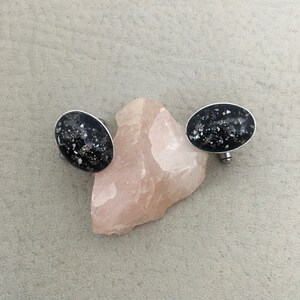 May include: Oval-shaped black earrings with a silver border, speckled with white and iridescent fragments. The earrings are displayed on a light pink stone against a neutral gray background. The earrings have a post backing.