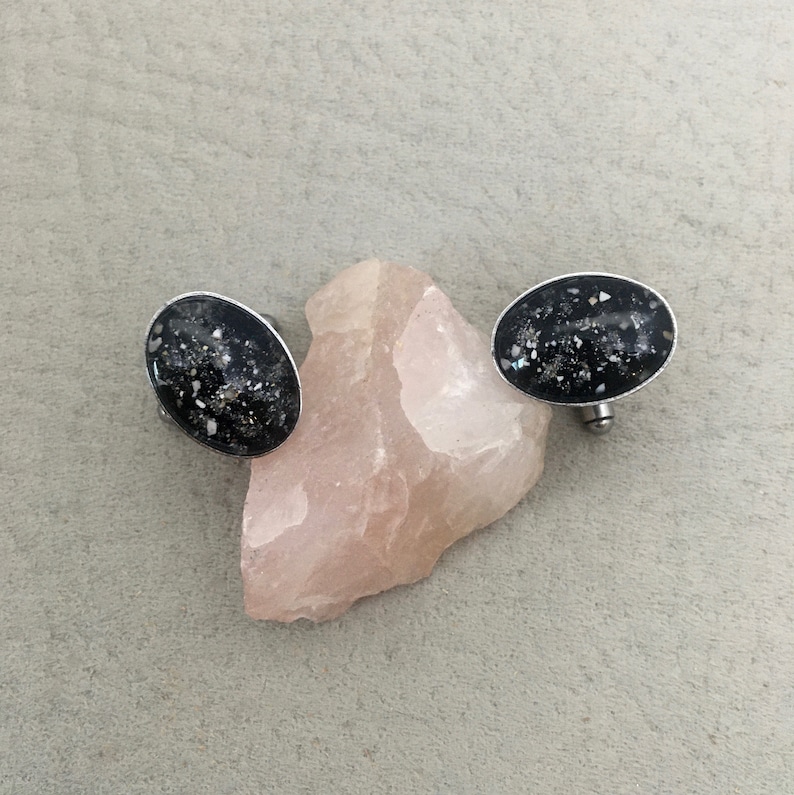 May include: Oval-shaped black earrings with a silver border, speckled with white and iridescent fragments. The earrings are displayed on a light pink stone against a neutral gray background. The earrings have a post backing.