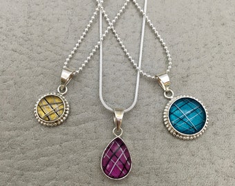 Puede incluir: Tres collares de plata con diferentes colgantes. Los colgantes son redondos con un diseño estampado en dorado, azul verdoso y rosa.