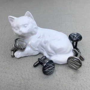 May include: A collection of cufflinks with a geometric design. The cufflinks have a silver-tone metal base and a round, black or silver face with a white, linear pattern. The cufflinks are arranged near a white ceramic cat figurine.