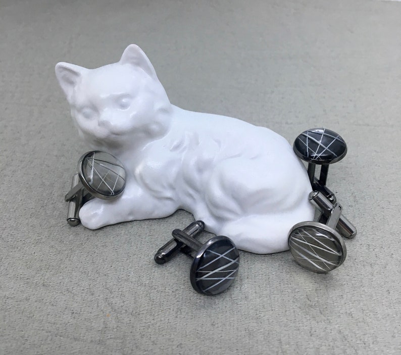 May include: A collection of cufflinks with a geometric design. The cufflinks have a silver-tone metal base and a round, black or silver face with a white, linear pattern. The cufflinks are arranged near a white ceramic cat figurine.