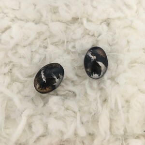 May include: Two oval-shaped earrings with a black, silver, and brown design. The earrings have a silver border and are set against a white, fluffy background. The earrings have a unique, abstract pattern.