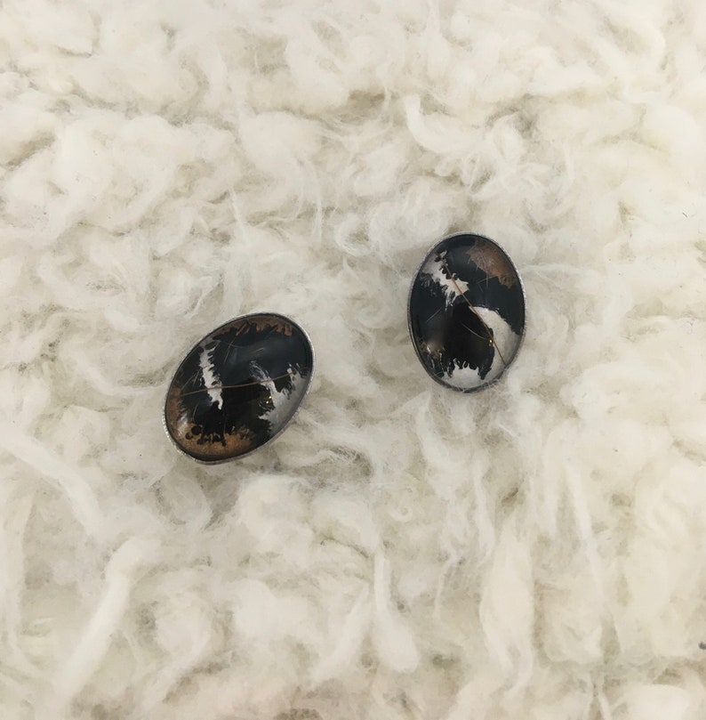 May include: Two oval-shaped earrings with a black, silver, and brown design. The earrings have a silver border and are set against a white, fluffy background. The earrings have a unique, abstract pattern.