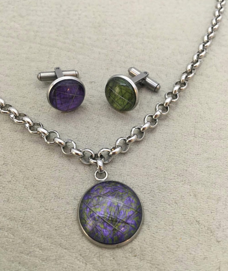 May include: A silver-toned necklace with a matching pendant and a pair of cufflinks. The pendant and cufflinks feature a circular design with a purple and green pattern. The chain and cufflink hardware are a shiny silver color.