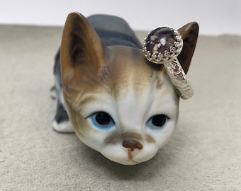 May include: A sterling silver ring with a round, ornate setting. The setting holds a dark purple stone with flecks of gold and white. The ring is displayed on a ceramic cat figurine.