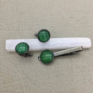 May include: A set of cufflinks and a tie clip with a matching design. The accessories feature a green and black geometric pattern set in a silver-tone metal. The cufflinks and tie clip are displayed on a white rectangular object.