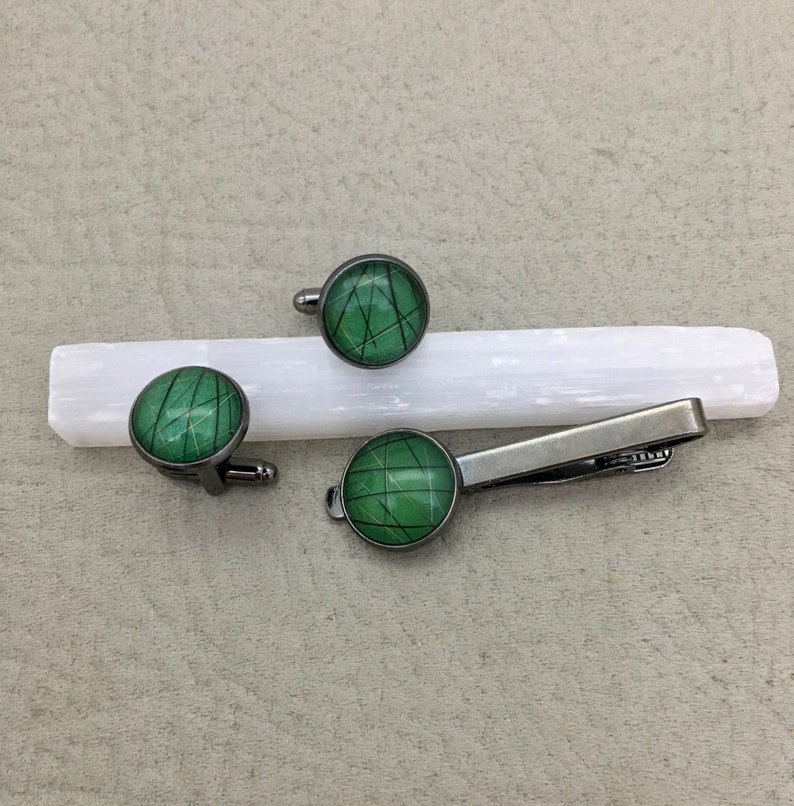 May include: A set of cufflinks and a tie clip with a matching design. The accessories feature a green and black geometric pattern set in a silver-tone metal. The cufflinks and tie clip are displayed on a white rectangular object.