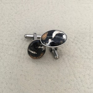 May include: A pair of oval cufflinks with a black, white, and gold marbled design. The cufflinks have a silver-colored metal backing and post. The design is reminiscent of natural stone or a geological pattern.