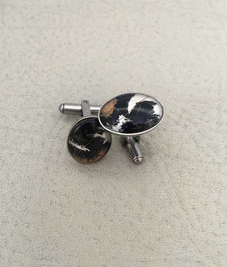 May include: A pair of oval cufflinks with a black, white, and gold marbled design. The cufflinks have a silver-colored metal backing and post. The design is reminiscent of natural stone or a geological pattern.