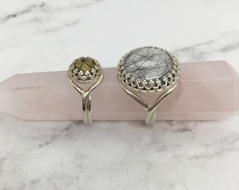 May include: Two silver rings with different colored stones. The larger ring has a white stone with black veins, and the smaller ring has a yellow stone. The rings are displayed on a pink crystal.