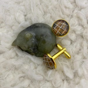 May include: Gold-toned cufflinks with a brown and white grid pattern. The cufflinks are round with a gold border and a T-bar closure. The background is a soft, white, fluffy material.