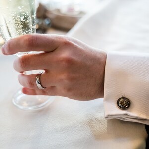 May include: A close-up of a person's hand holding a champagne flute. The person is wearing a silver ring and a white dress shirt with a black suit jacket. The cufflink is a dark metal with a circular design.