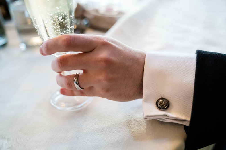 May include: A close-up of a person's hand holding a champagne flute. The person is wearing a silver ring and a white dress shirt with a black suit jacket. The cufflink is a dark metal with a circular design.
