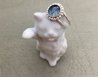 May include: A silver ring with a blue stone set in a beaded bezel. The ring is on a white ceramic cat figurine.