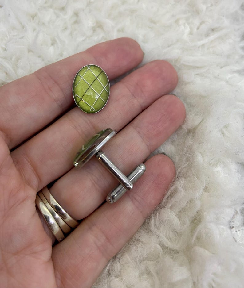 May include: A pair of cufflinks with a lime green oval design and a silver grid pattern. The cufflinks have a silver metal backing and a post for securing them to a shirt cuff. The design is repeated on the oval button.