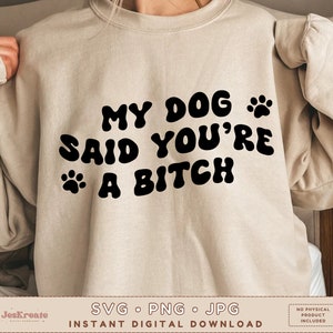 Op de afbeelding: Een beige sweatshirt met zwarte tekst die "My Dog Said You're A Bitch" zegt met een poot afdruk grafiek aan beide kanten van de tekst.