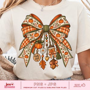 Op de afbeelding: Een crèmekleurig t-shirt met een grote afbeelding van een decoratieve strik met oranje pompoenen, ruitpatronen en herfstbladeren. Hangen aan de strik zijn bedels zoals een koffiebeker, een eikel, een madeliefje en een label met de tekst hello fall.