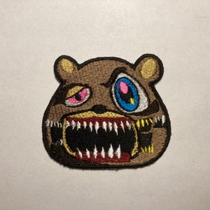 May include: Embroidered patch featuring a brown bear with a menacing expression. The bear has a wide open mouth with sharp white teeth, a pink and red eye, and a blue and yellow eye. The patch is likely for clothing or accessories.