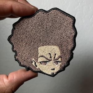 May include: Embroidered patch of a cartoon character with a brown afro, furrowed brows, and a serious expression.