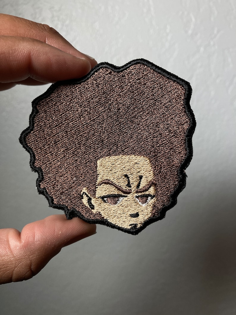 May include: Embroidered patch of a cartoon character with a brown afro, furrowed brows, and a serious expression.