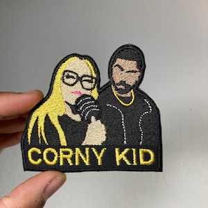 May include: A black embroidered patch with a yellow outline featuring a cartoon image of a woman with blonde hair holding a microphone and a man with a beard and a gold chain. The text "CORNY KID" is embroidered in yellow below the cartoon figures.