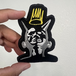 May include: A black embroidered patch featuring a stylized grey silhouette of a head with pointed ears, topped with a bright yellow crown icon. The design is outlined in black thread, creating a distinct shape against the white background.