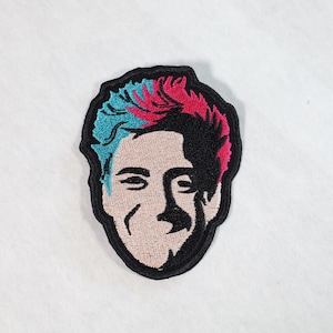 May include: Embroidered patch featuring a cartoon portrait of a person with blue and pink hair. The person has a black outline and a light skin tone.