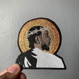 May include: Embroidered patch featuring a black and brown profile of a person wearing a crown of thorns, with a gold halo behind them. The patch is on a white background.