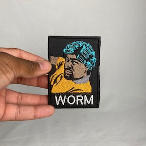 May include: Embroidered patch featuring a stylized portrait of a person with blue curlers and a yellow shirt. The word "WORM" is embroidered in white letters at the bottom of the black patch.