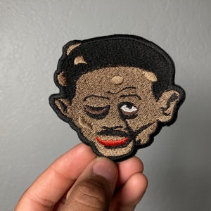 May include: A black and brown embroidered patch of a cartoon man with a black afro, winking, and a red mouth.