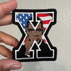 May include: An embroidered patch shaped like the letter X with a black border and white outline. The design features a portrait of a person with glasses, set against a background of the American flag with blue stars on the left and red stripes on the right.