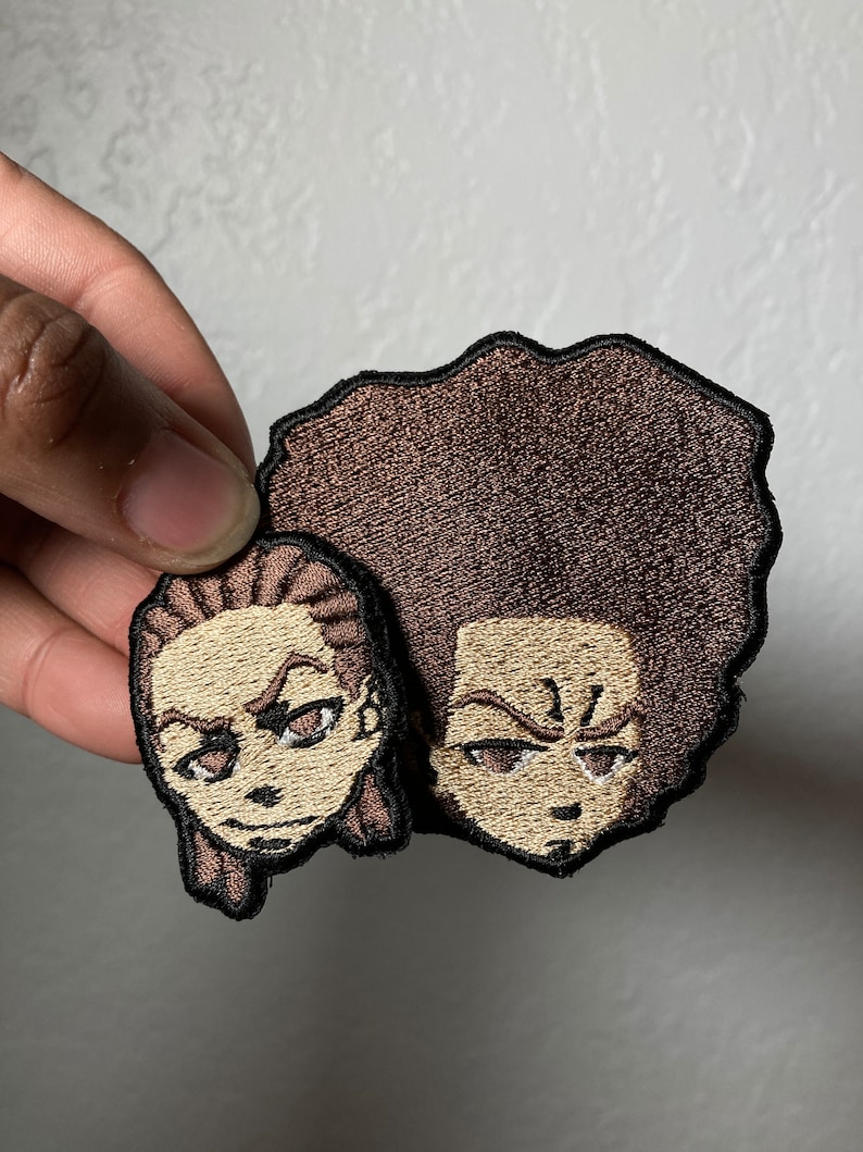 May include: Two embroidered patches featuring cartoon characters with brown hair and brown skin. The characters have a serious expression and are looking to the right. The patches are brown and beige with black outlines.