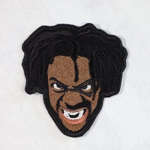 May include: Embroidered patch featuring a cartoon face with black hair, brown skin, and a menacing expression. The face is outlined in black and the eyes are white with black pupils.