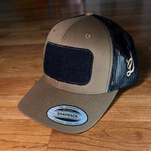 May include: A brown and black snapback hat with a black patch on the front. The hat has a mesh back and a curved brim. The text "THE AUTHENTIC SNAPBACK YUPOONG SINCE 1974" is on the brim.