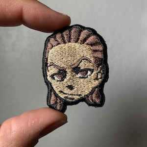 May include: Embroidered patch featuring a cartoon face with brown hair, brown eyes, and a light brown skin tone. The patch has a black outline.
