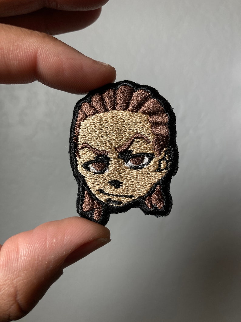 May include: Embroidered patch featuring a cartoon face with brown hair, brown eyes, and a light brown skin tone. The patch has a black outline.