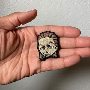 May include: A brown and beige embroidered patch of a cartoon character with brown hair and a serious expression.