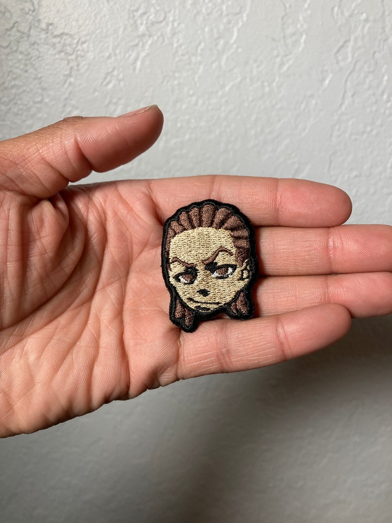 May include: A brown and beige embroidered patch of a cartoon character with brown hair and a serious expression.