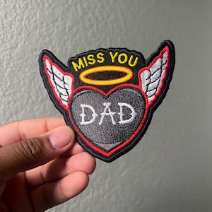 May include: Embroidered patch with a heart-shaped design. The patch features the words "MISS YOU DAD" in white lettering, with a yellow halo above the heart and white wings on either side. The patch has a black background with red trim.