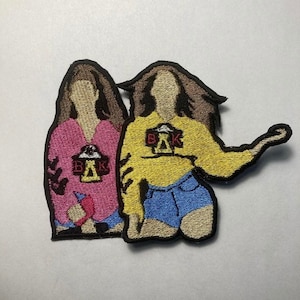 May include: Two embroidered patches of women wearing different outfits. The woman on the left is wearing a pink shirt and the woman on the right is wearing a yellow shirt and blue denim shorts. Both women have brown hair and are wearing patches with the letters "BAK" and a symbol.