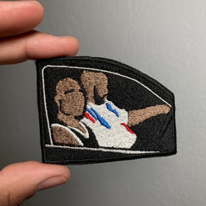 May include: Black and white embroidered patch featuring two people in a car. The person in the driver's seat is wearing a white shirt with blue and red accents. The passenger is wearing a brown shirt. The patch has a white outline.