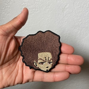 May include: A brown and black embroidered patch of a cartoon character with a large afro hairstyle. The character has a serious expression with a furrowed brow and a small frown.
