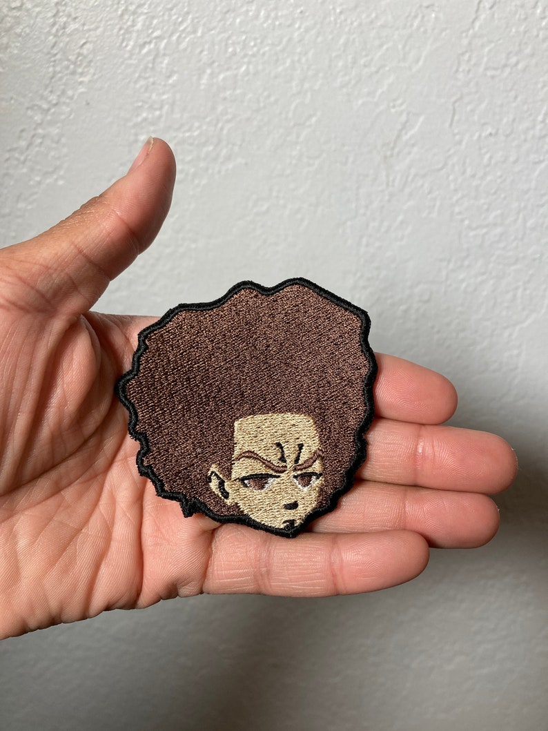 May include: A brown and black embroidered patch of a cartoon character with a large afro hairstyle. The character has a serious expression with a furrowed brow and a small frown.